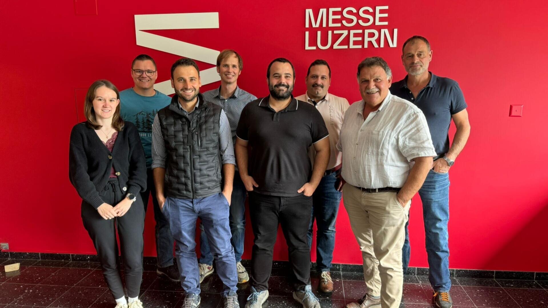 Acht Personen stehen lächelnd vor einer leuchtend roten Wand mit der weißen Aufschrift MESSE LUZERN. Sie wirken lässig und freundlich, posieren als Gruppe in einem Innenraum.
