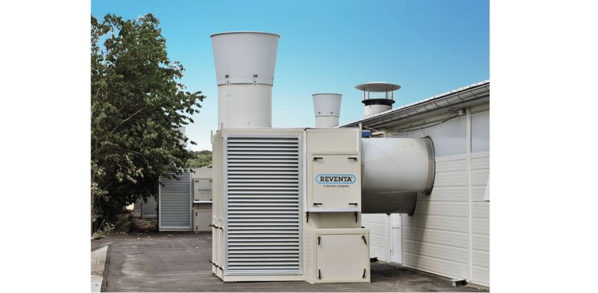 Große industrielle Lüftungsanlagen mit der Aufschrift Reventa sind an der Seite eines Gebäudes angebracht, und hohe Abluftkamine ragen vor einem klaren blauen Himmel in die Höhe, während im Hintergrund ein Baum zu sehen ist.