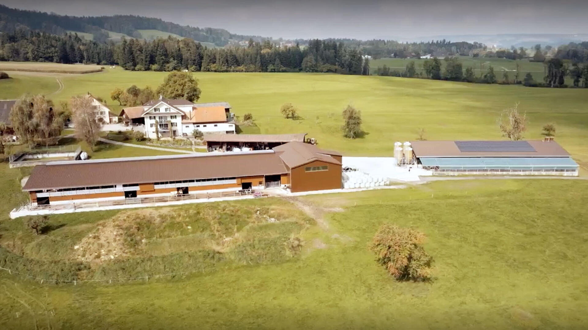Luftaufnahme eines ländlichen Bauernhofs mit mehreren großen braunen Gebäuden, Silos und einem Gewächshaus mit Sonnenkollektoren, umgeben von grünen Feldern und verstreuten Bäumen. Im Hintergrund sind Häuser und bewaldete Hügel zu sehen.
