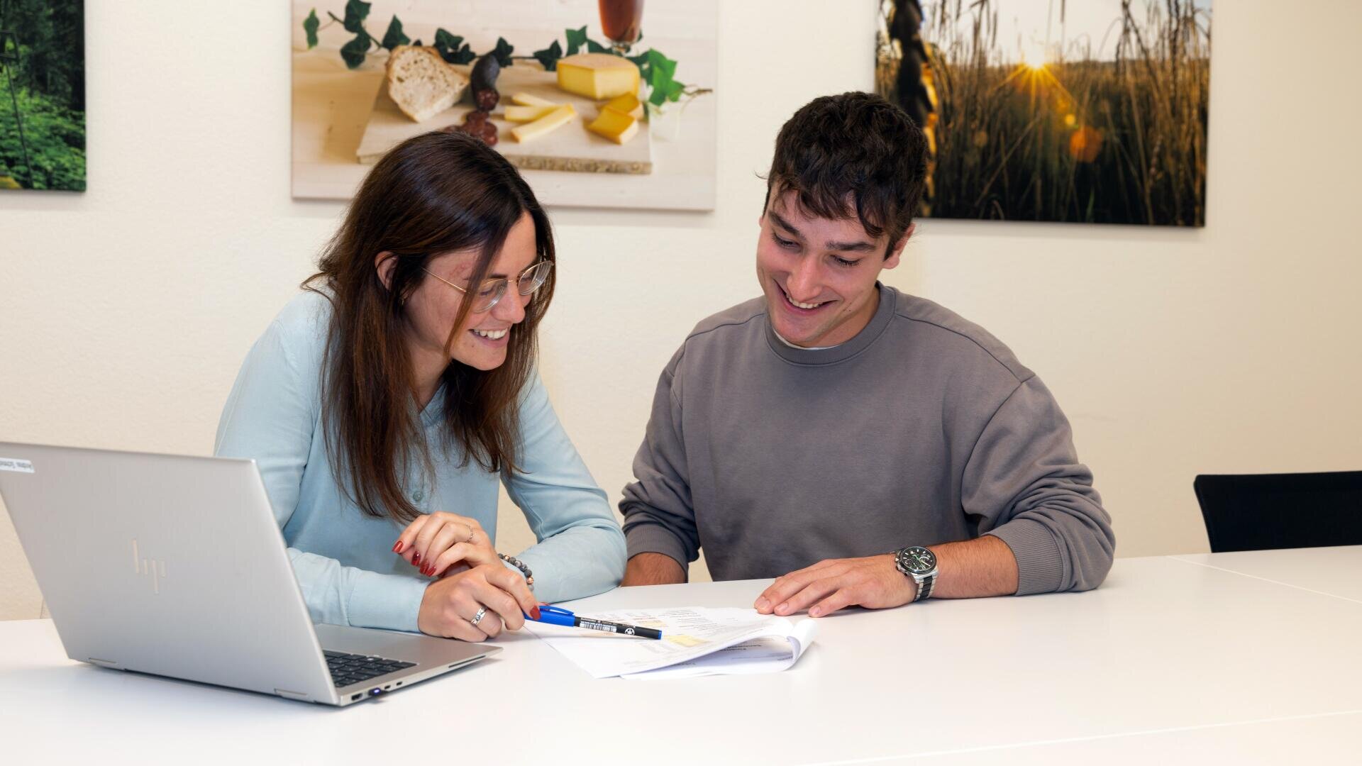 Zwei Personen sitzen an einem weißen Tisch mit einem Laptop und arbeiten lächelnd an Dokumenten. Bunte Fotos schmücken die Wand hinter ihnen, darunter Bilder von Lebensmitteln und einem Feld.