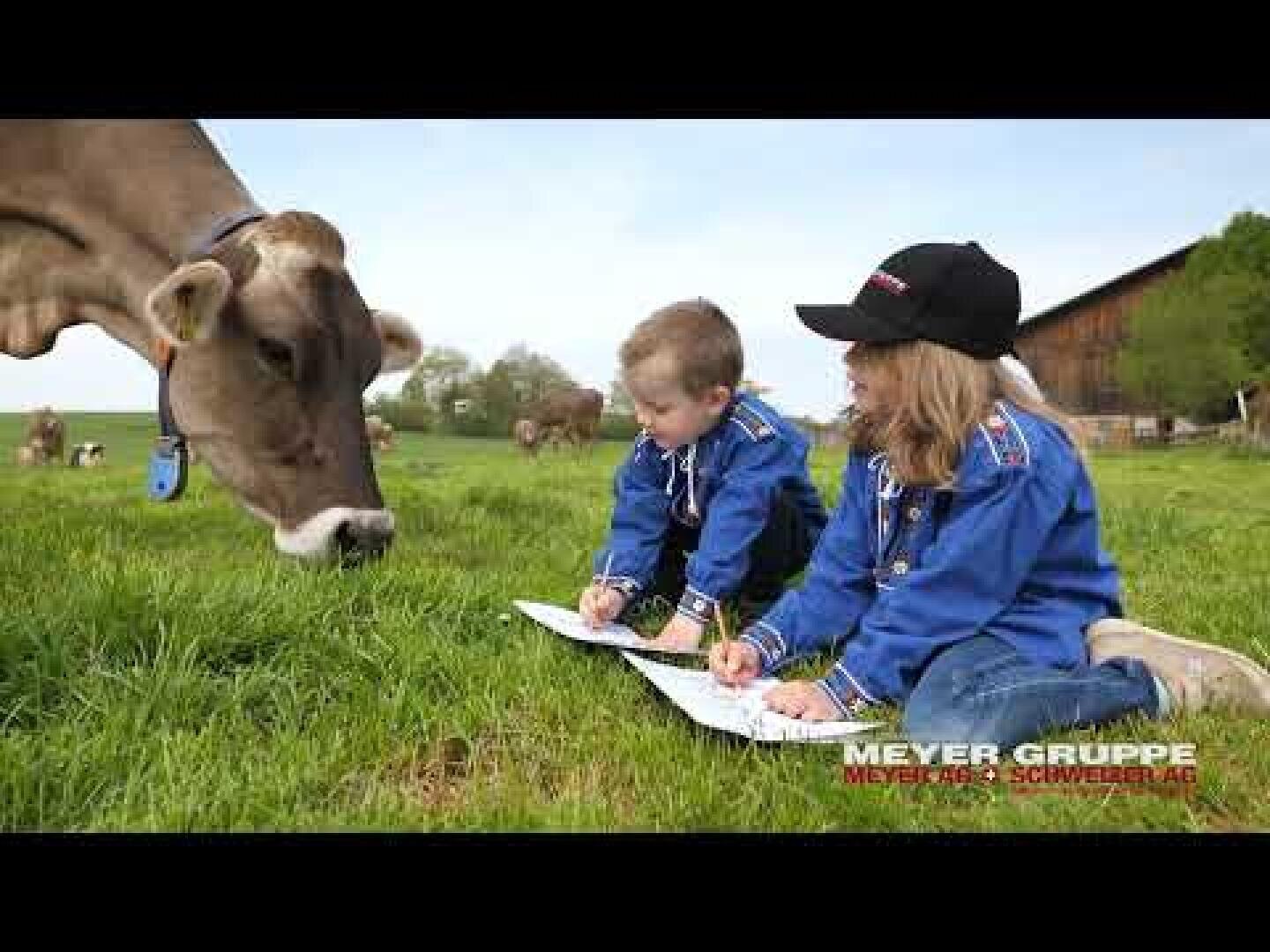 Zwei Kinder in blauen Jacken sitzen im Gras und zeichnen auf Papier, während in der Nähe eine braune Kuh grast. Im Hintergrund sind Kühe auf einer Wiese in der Nähe eines Stalls bei klarem Himmel zu sehen.