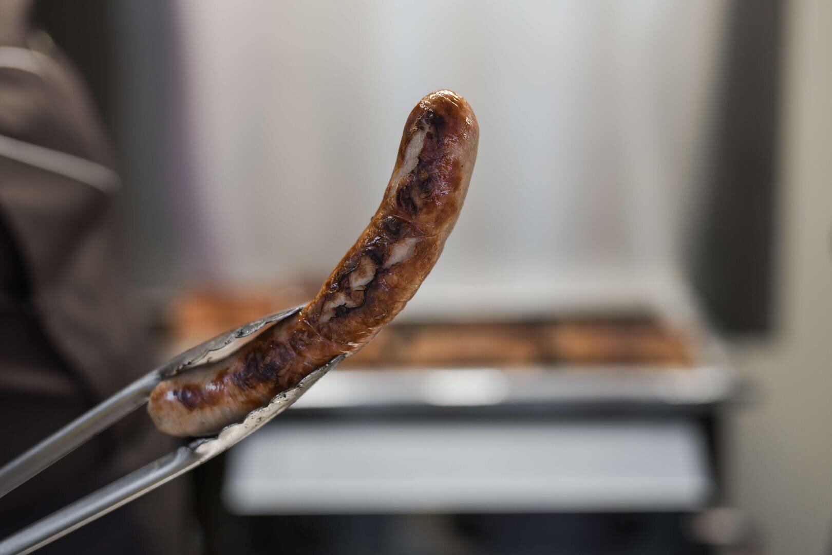 Eine Nahaufnahme einer gegrillten Wurst, die mit einer Metallzange gehalten wird, mit einem unscharfen Hintergrund, der eine Küche oder einen Bereich für die Zubereitung von Lebensmitteln zu sein scheint.