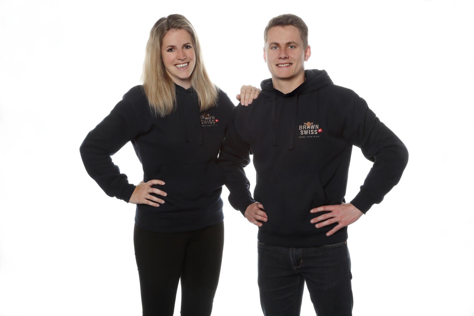 Zwei Personen in passenden marineblauen Kapuzenpullovern mit "Brown Swiss"-Logo posieren lächelnd in die Kamera vor einem schlichten weißen Hintergrund. Eine Person hat den Arm um die Schulter der anderen gelegt, und beide haben die Hände in die Hüften gestemmt.