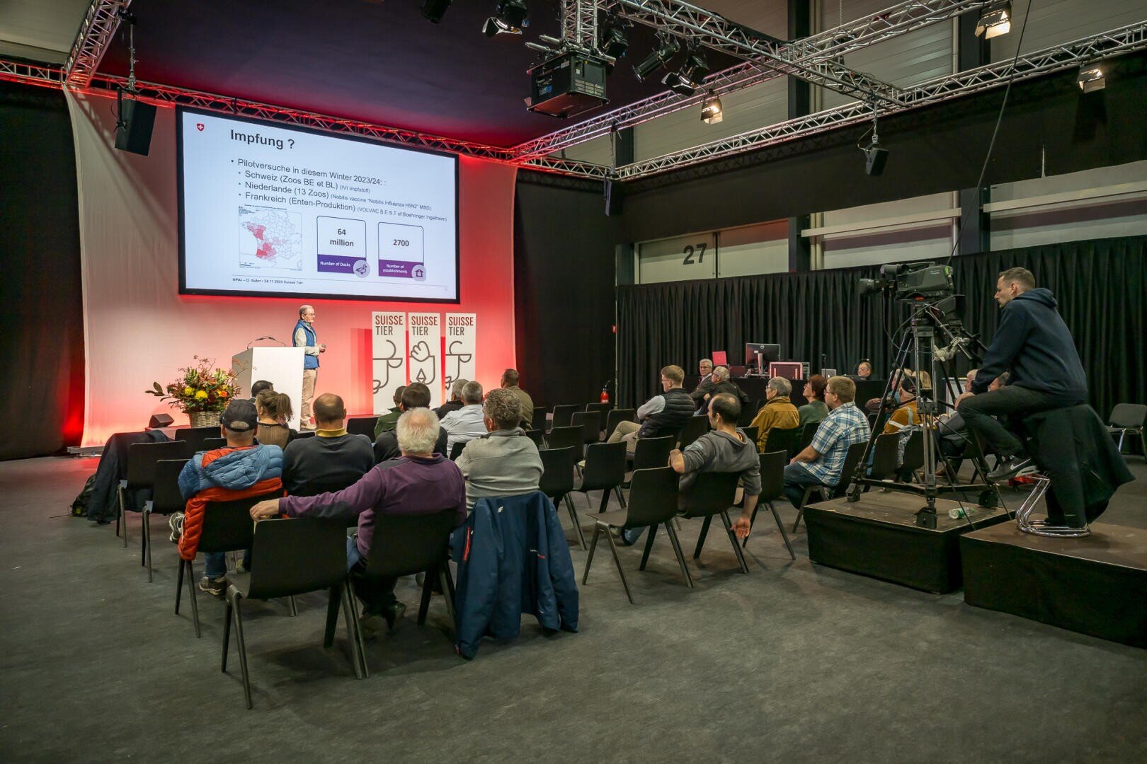 Ein Redner präsentiert einem sitzenden Publikum in einem Konferenzraum Informationen über Impfungen auf einem großen Bildschirm. Ein Kameramann zeichnet die Veranstaltung auf, und Blumen schmücken die Bühne.
