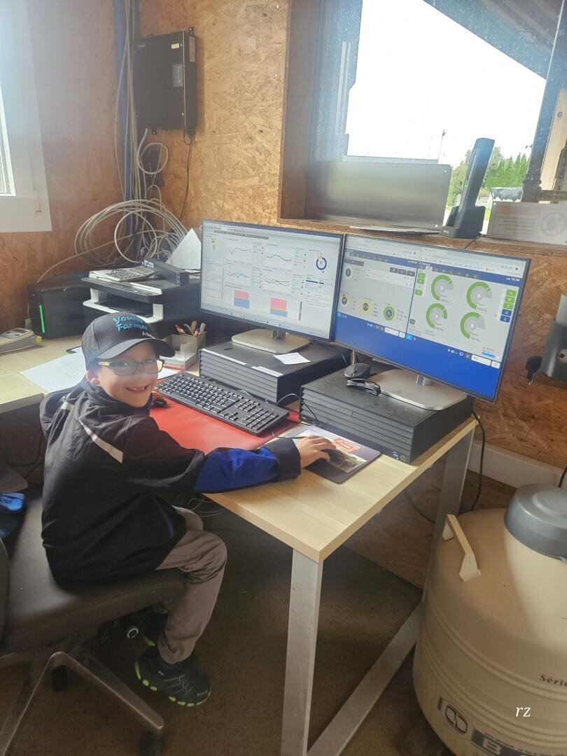 Ein junger Mann mit Brille, Mütze und Jacke sitzt an einem Schreibtisch vor zwei Computermonitoren, auf denen Diagramme und Grafiken angezeigt werden. Er lächelt in die Kamera und hat die Hand auf einer Computermaus in einem rustikalen Büro.
