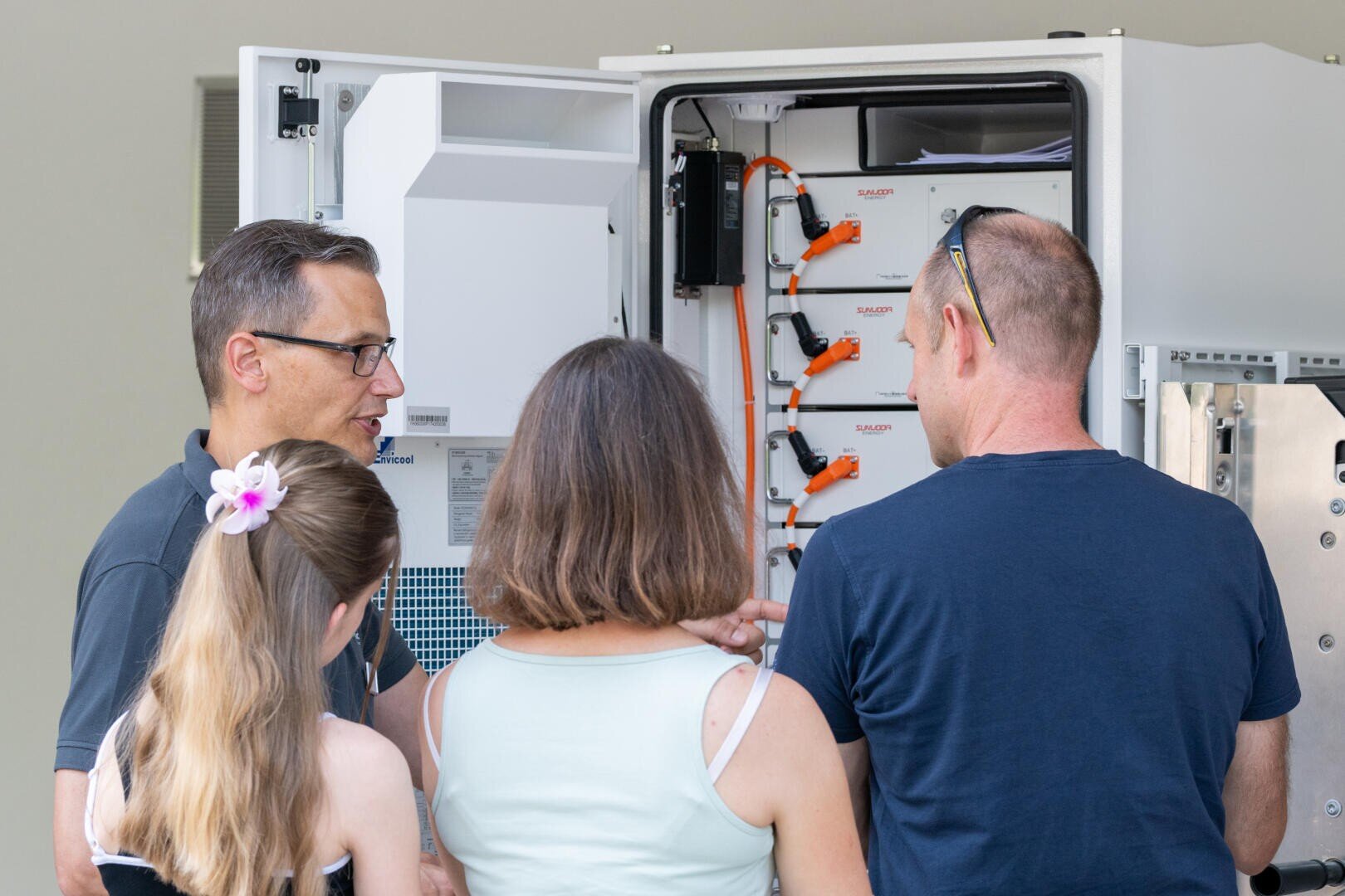 Drei Erwachsene und ein Kind stehen vor einem offenen Schaltschrank. Ein Mann erklärt oder bespricht die darin befindlichen Geräte mit sichtbaren Kabeln und beschrifteten Bauteilen. Die Gruppe scheint gemeinsam zu lernen oder Fehler zu beheben.