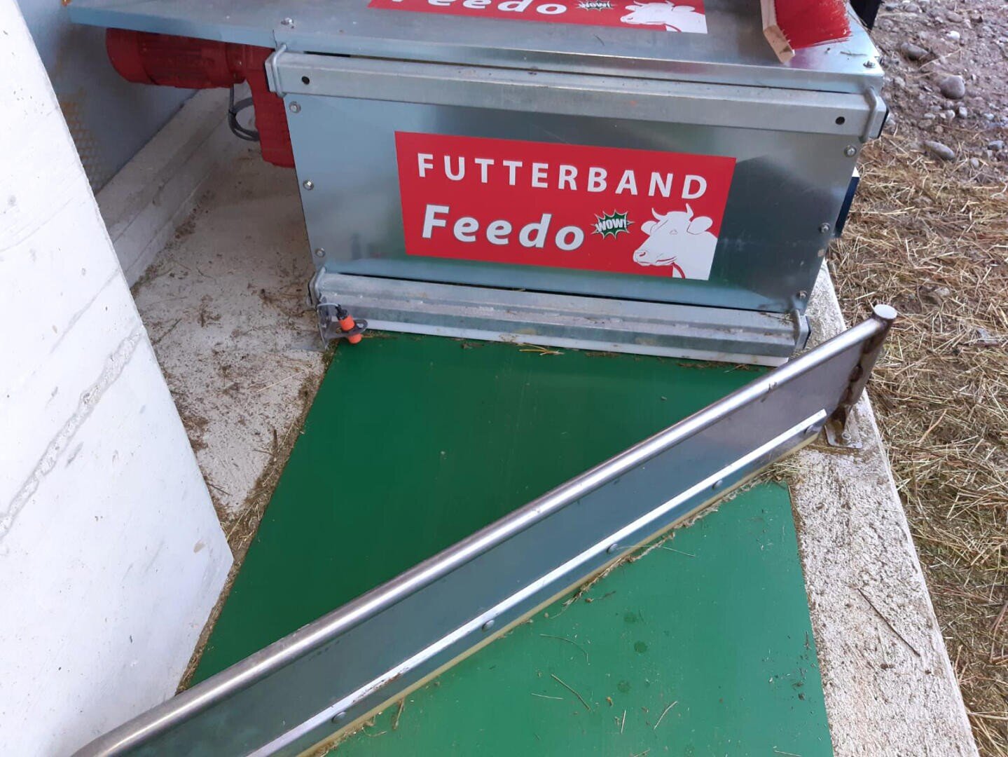 Eine Metallmaschine mit der Aufschrift FUTTERBAND Feedo und einem Cartoon-Kuhkopf steht auf einer grünen Matte neben einer Metallschiene, auf der etwas Heu verstreut ist.