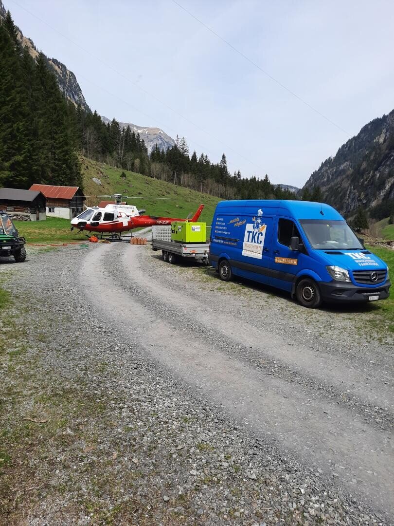 Ein blauer TKC-Kleintransporter parkt auf einer Schotterstraße neben einer grünen Wiese; dahinter stehen ein rot-weißer Hubschrauber, ein Anhänger und grüne Geräte, im Hintergrund bewaldete Hügel und ein Berg.