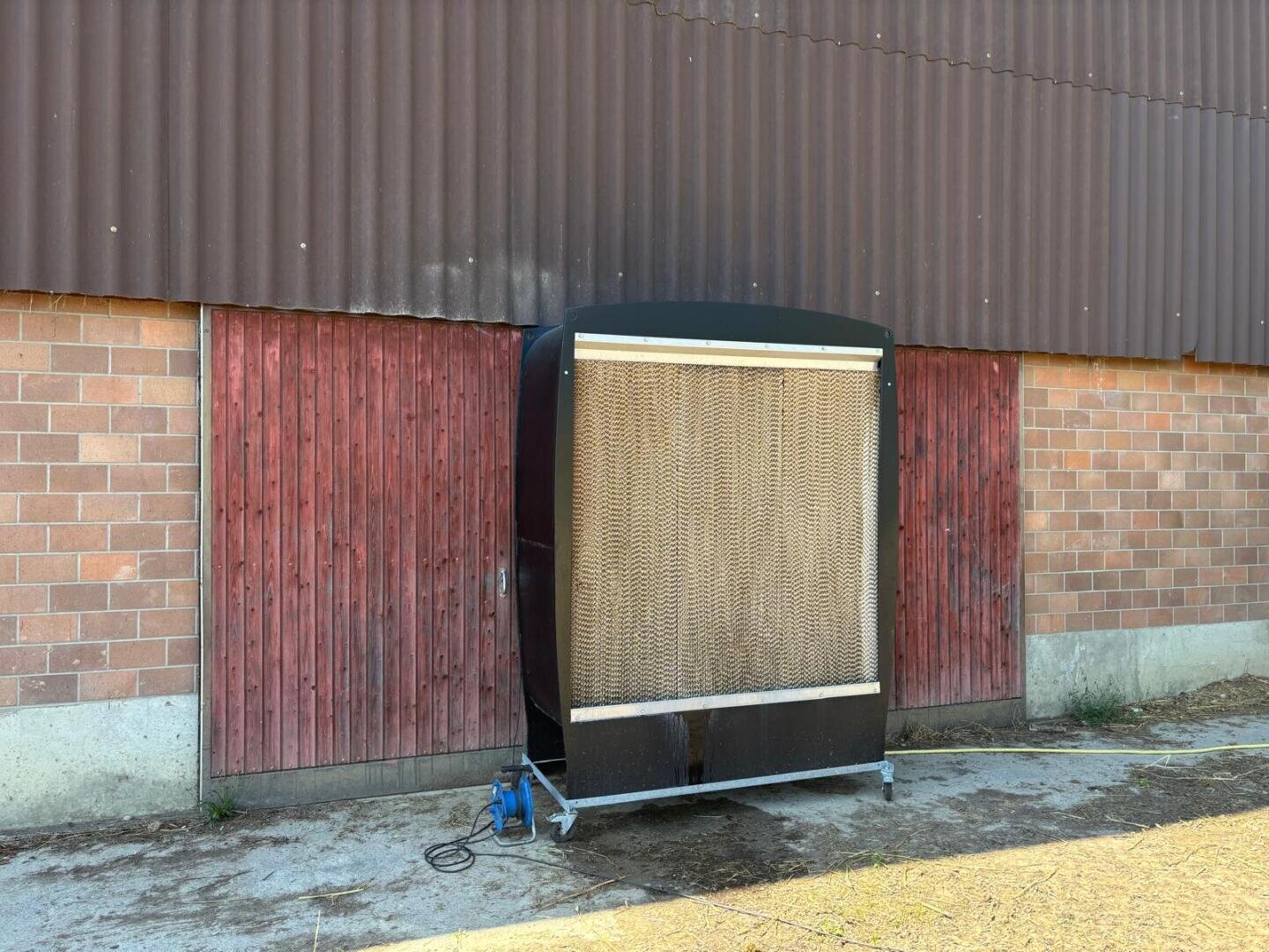 Ein großes schwarzes Industriegerät mit einer Gitterfront steht auf Rädern vor roten Holztüren an einem Backsteingebäude. Neben dem Gerät stehen ein blauer Kanister und ein Schlauch auf dem Boden.