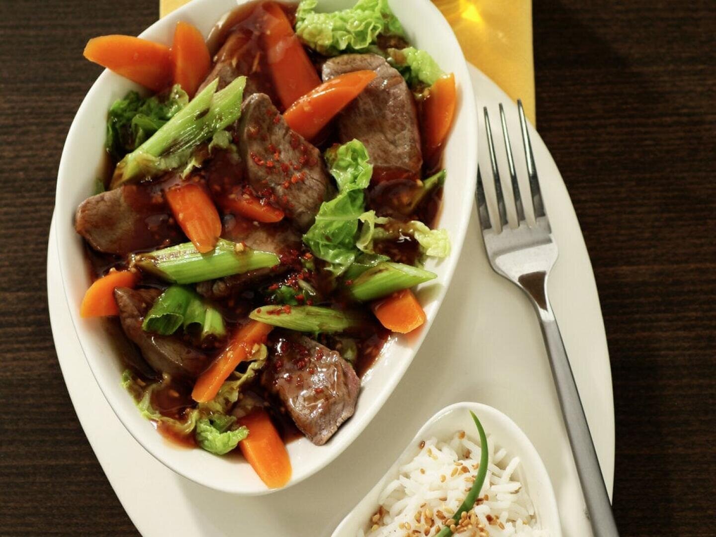 Eine weiße, ovale Schale mit gebratenem Rindfleisch, Karotten, grünen Zwiebeln und Kohl in einer braunen Soße, die auf einem Teller neben einer kleinen Schüssel mit weißem Reis steht, der mit Sesam und einer grünen Beilage garniert ist. Eine Gabel liegt daneben.