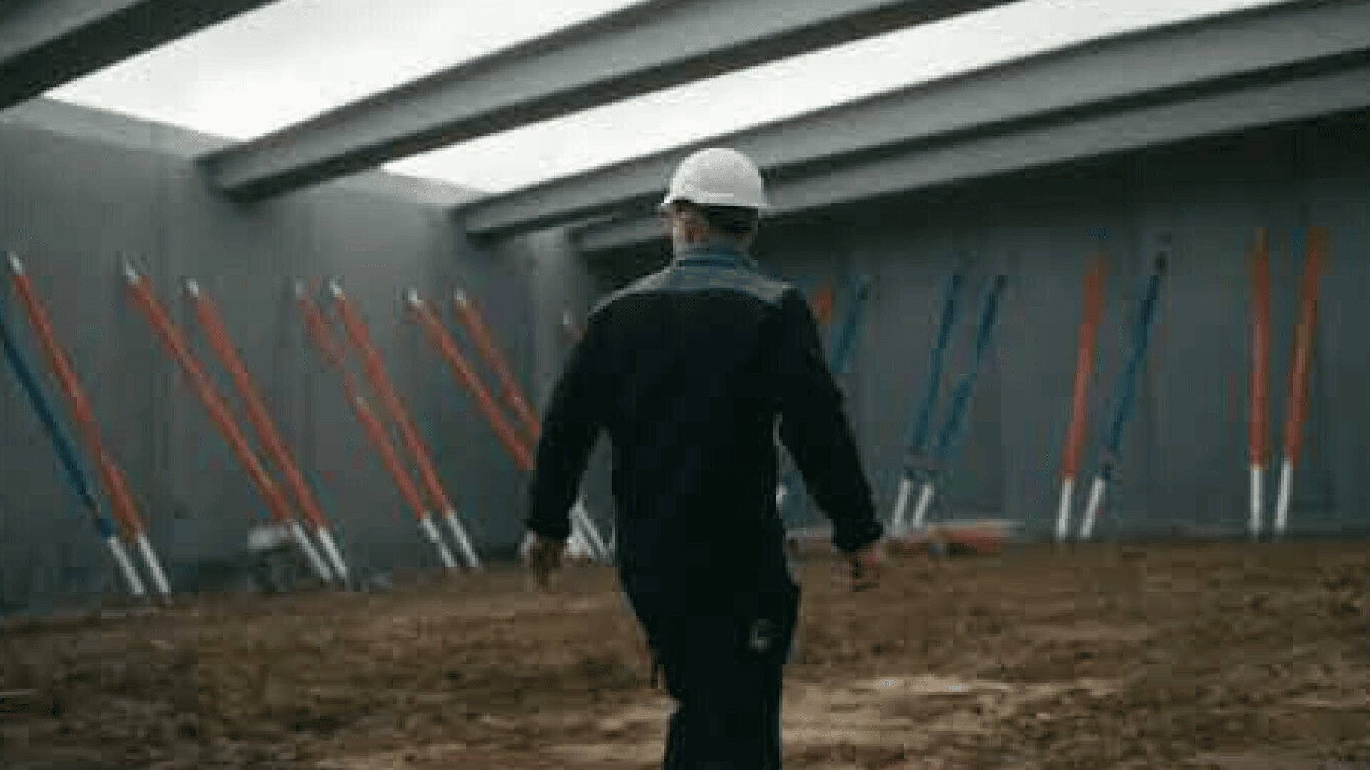 Ein Bauarbeiter, der einen weißen Schutzhelm und dunkle Kleidung trägt, geht in ein teilweise gebautes Gebäude mit freiliegenden Balken und Stützen entlang der Betonwände und steht auf einem schmutzigen Boden.