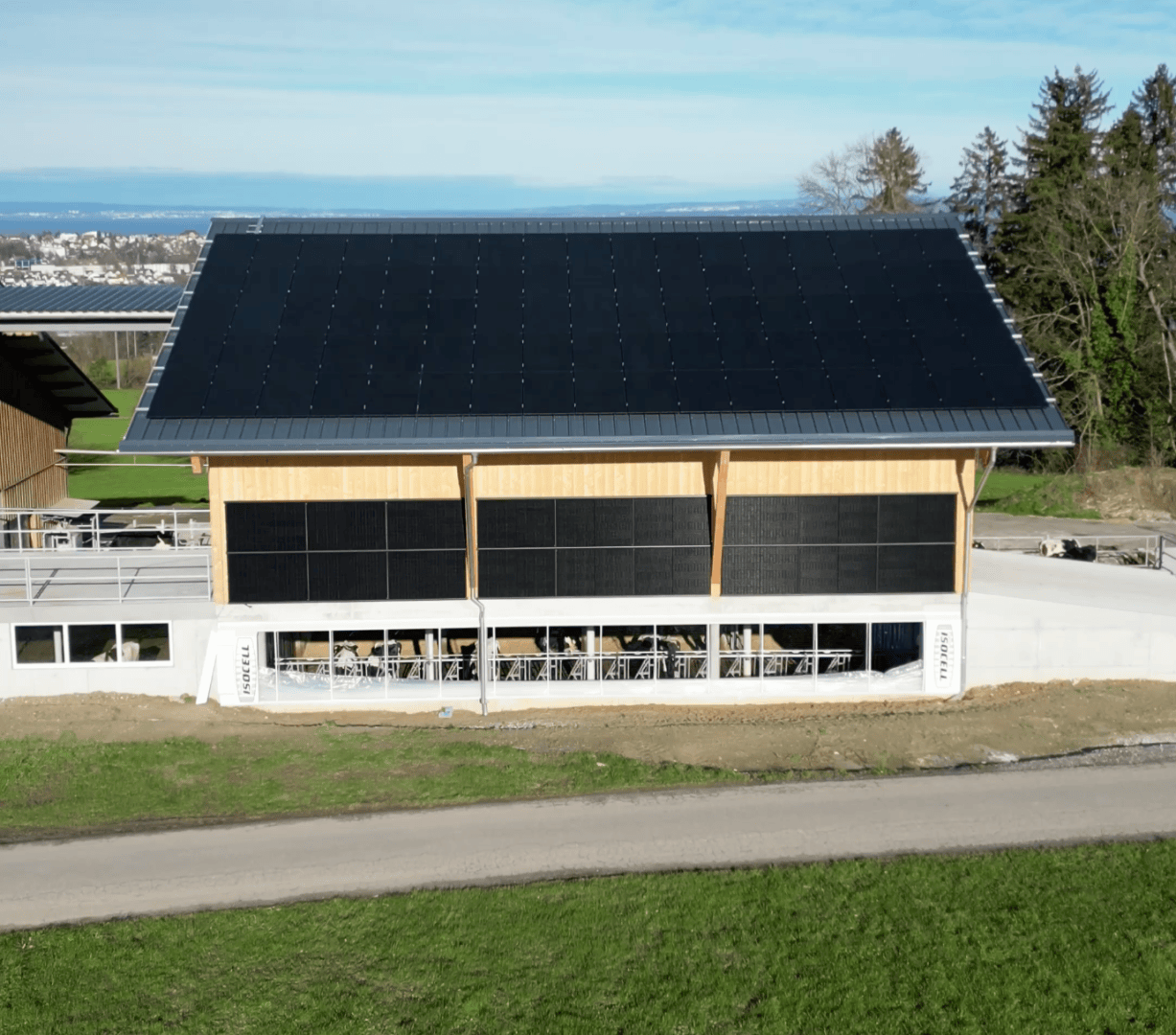 Eine moderne Scheune mit einem großen Schrägdach und Außenwänden, die mit schwarzen Solarpaneelen bedeckt sind, umgeben von Gras, mit einem klaren blauen Himmel und entfernten Gebäuden im Hintergrund.