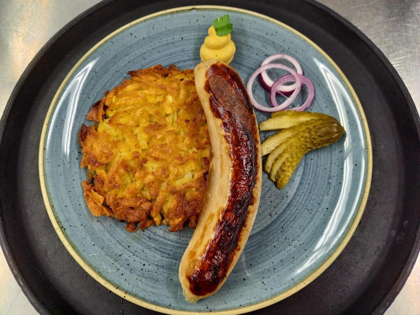 Ein blauer Teller mit einer gebratenen Wurst, einem goldenen Kartoffelpuffer, in Scheiben geschnittenen Essiggurken, roten Zwiebelringen, einem Klecks Senf und einem Blatt Petersilie als Garnitur, alles auf einem schwarzen Tablett.