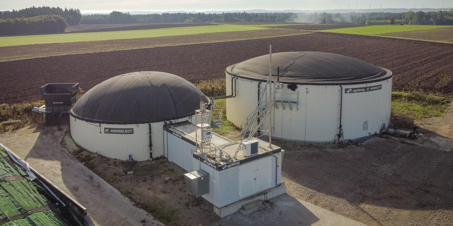 Luftaufnahme von zwei großen, runden Biogastanks mit schwarzen Kuppeldächern auf einem Bauernhof, umgeben von Feldern und Bäumen, mit einem kleinen Kontrollgebäude und Geräten davor.