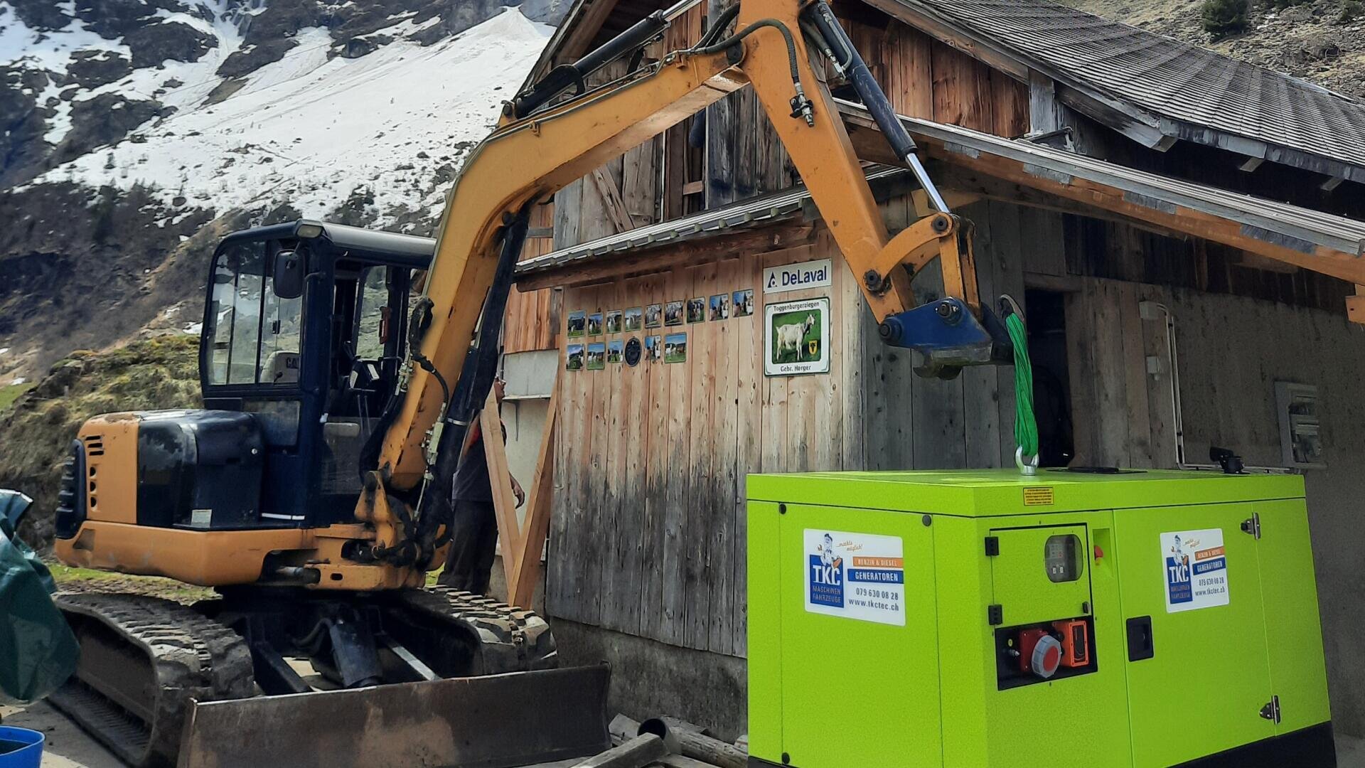 Ein gelber Bagger hebt einen leuchtend grünen Generator vor einem hölzernen Berggebäude mit schneebedeckten Gipfeln im Hintergrund an. An der Gebäudewand sind Schilder und Fotos angebracht.