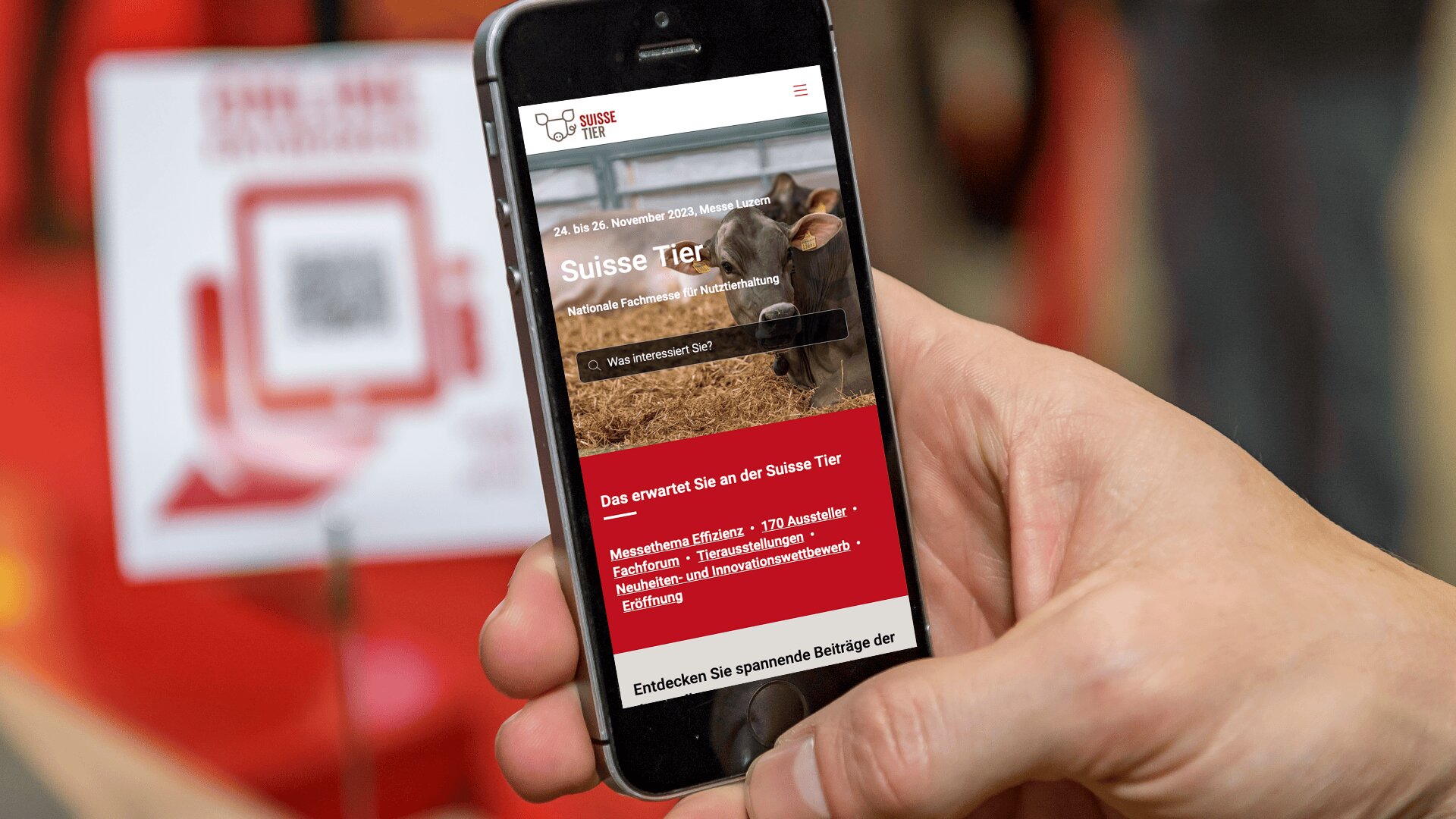 Eine Hand hält ein Smartphone, auf dem die Website von Suisse Tier mit dem Bild einer Kuh abgebildet ist. Im unscharfen Hintergrund sind ein QR-Code und eine rote Beschilderung zu sehen.