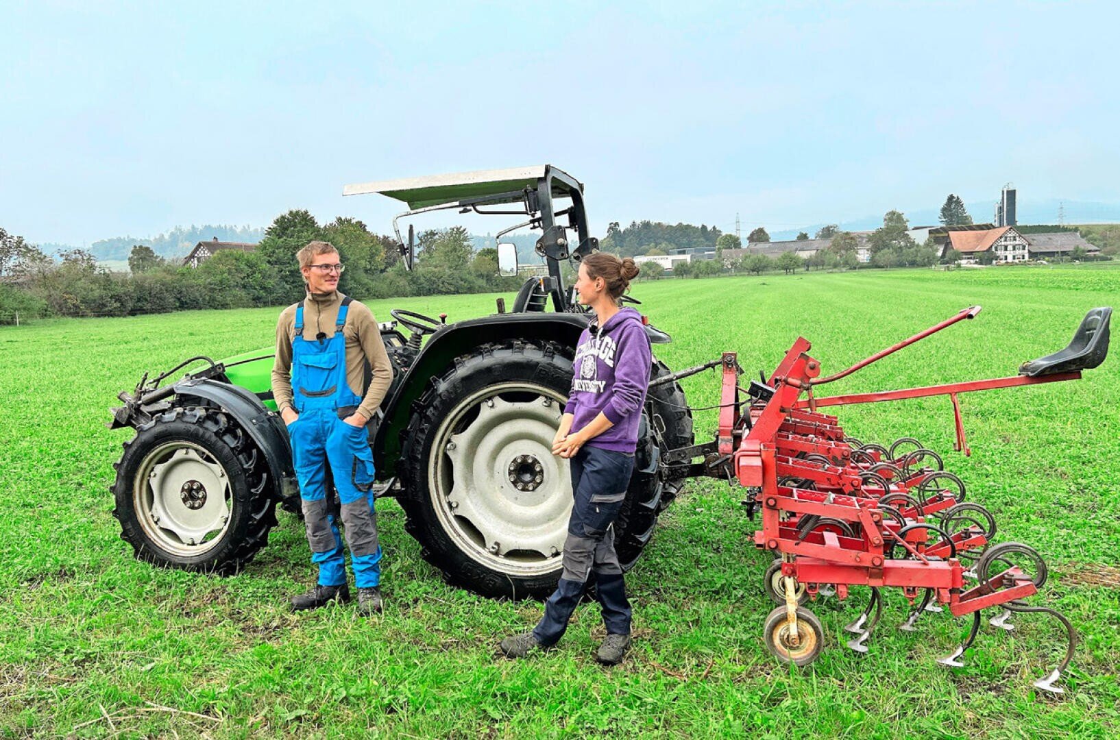 Zwei Personen stehen auf einem grünen Feld neben einem schwarzen Traktor, der an einem roten landwirtschaftlichen Gerät befestigt ist. Im Hintergrund sind Bäume und Gebäude unter einem bewölkten Himmel zu sehen.