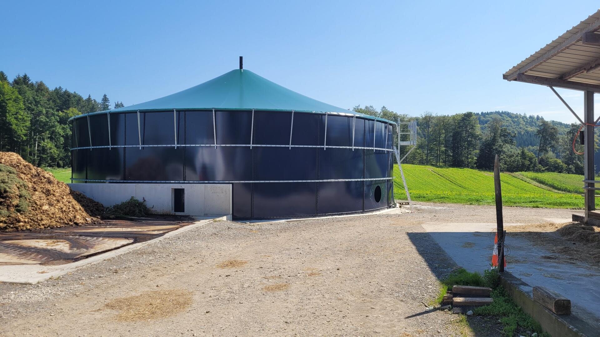 Ein großer, runder, dunkelblauer landwirtschaftlicher Lagertank mit einem grünen Kegeldach steht auf einem Bauernhof, umgeben von Feldern, Bäumen und einem klaren blauen Himmel. Eine kleine Leiter führt zu einer Öffnung an der Seite des Tanks.