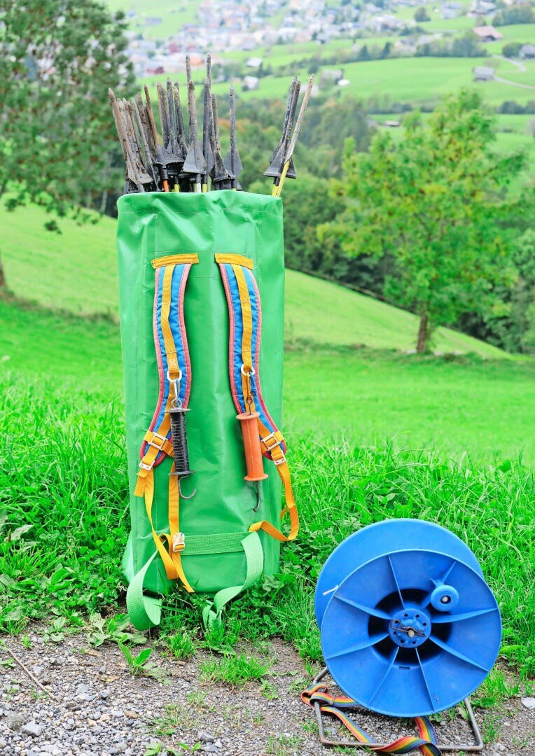 Ein grüner, mit Stöcken gefüllter Rucksack steht auf der Wiese neben einer blauen Kabeltrommel, während im Hintergrund grüne Hügel und ein Dorf zu sehen sind.