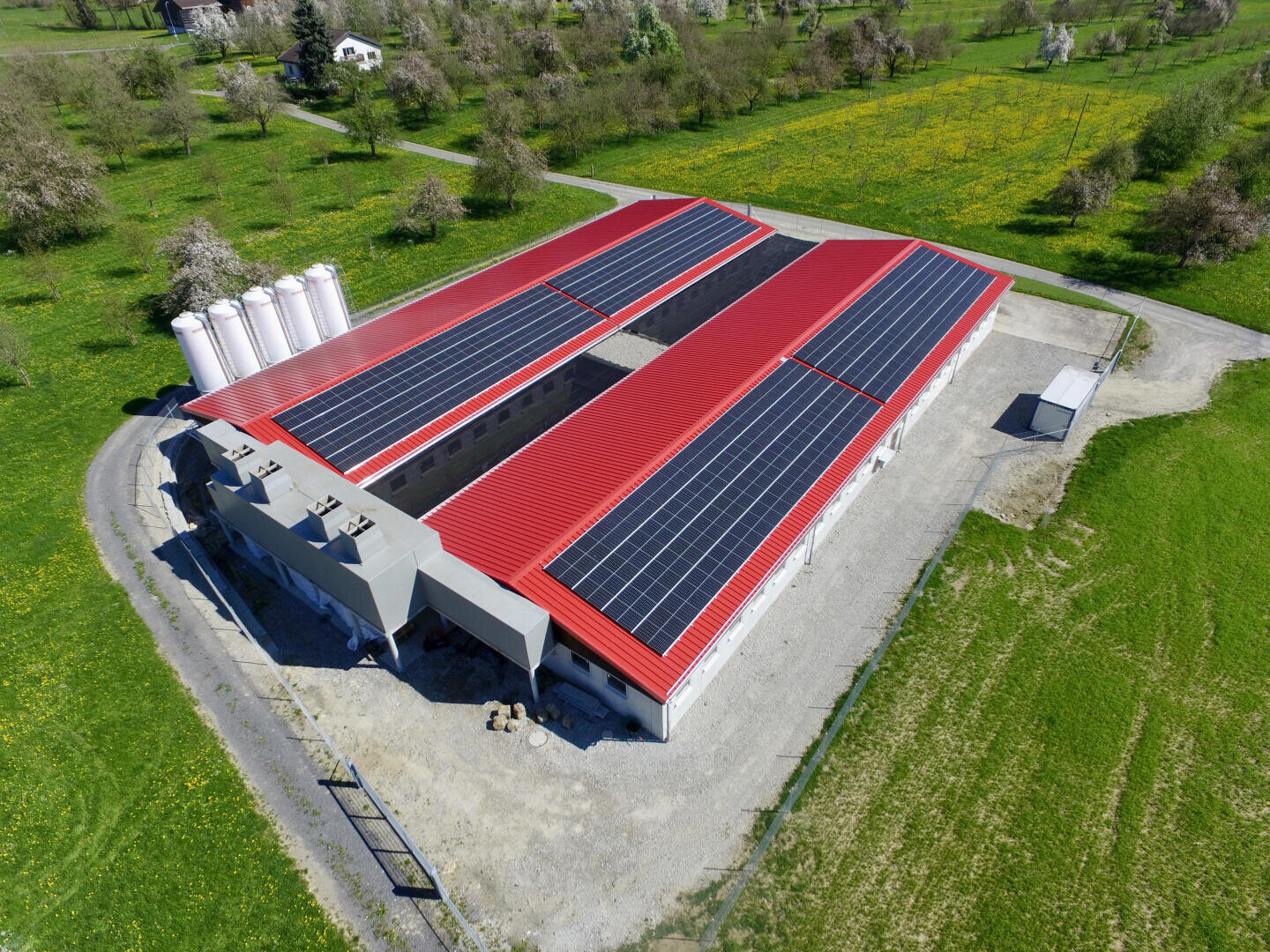 Luftaufnahme eines großen Gebäudes mit rotem Dach, das mit Solarzellen bedeckt ist, umgeben von grünem Gras und Bäumen, mit mehreren weißen zylindrischen Tanks neben dem Gebäude.