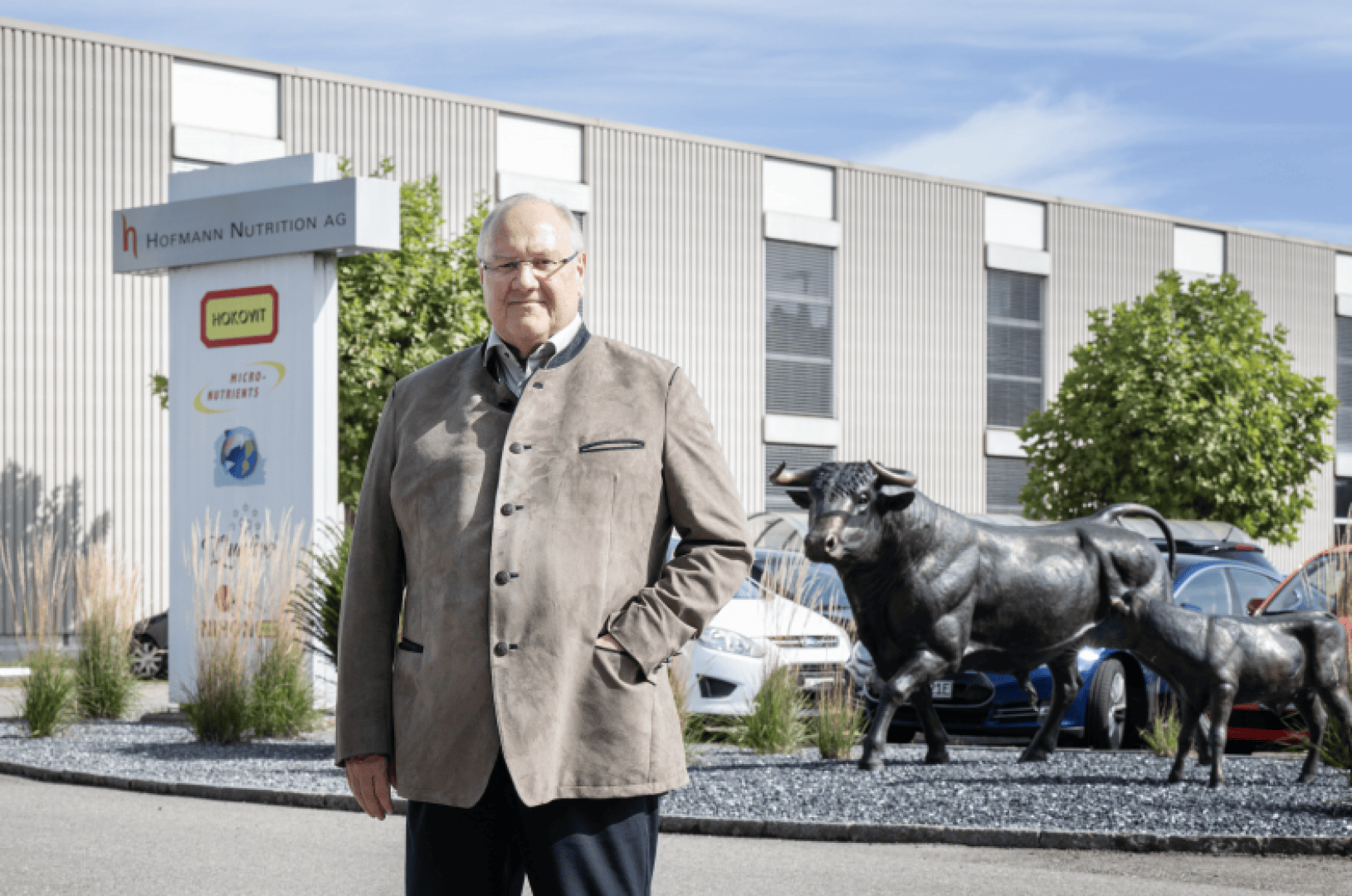 Ein älterer Mann in einer beigen Jacke steht vor einem modernen Gebäude mit einem Schild der Hofmann Nutrition AG. Hinter ihm stehen geparkte Autos, Grünflächen und eine große Stierskulptur.