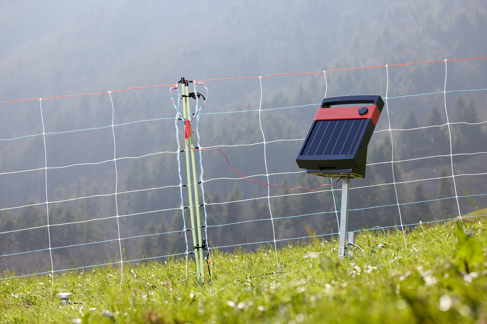 Ein tragbares solarbetriebenes Elektrozaungerät ist auf einer Wiese installiert. Im Hintergrund sind an einem dunstigen Tag ein Maschendrahtzaun und bewaldete Hügel zu sehen.