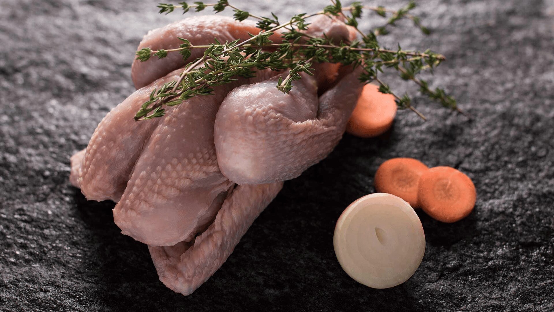 Rohe ganze Wachteln, garniert mit frischem Thymian, auf einer dunklen Steinplatte, daneben zwei Scheiben Karotten und eine Scheibe weiße Zwiebel.