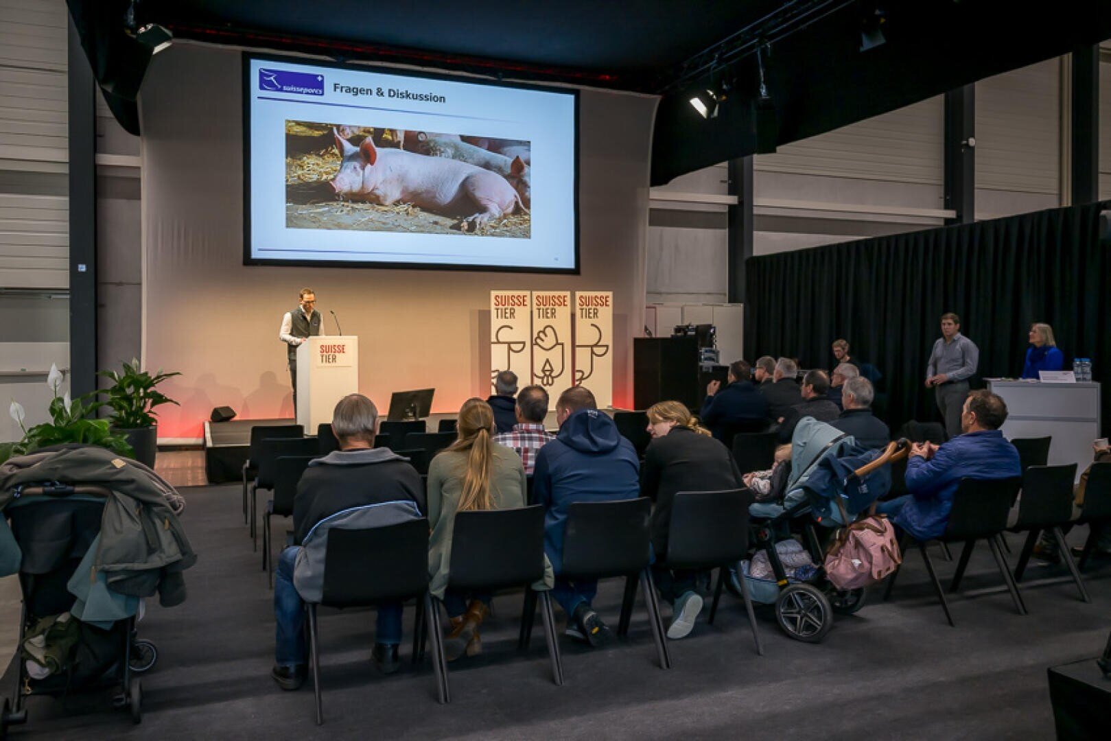 Ein Redner hält vor einem Publikum in einem Konferenzraum einen Vortrag und zeigt dabei auf einer Leinwand eine Folie mit Schweinen. Die Zuhörer sitzen mit dem Blick zur Bühne, und es sind Veranstaltungsbanner mit der Aufschrift „SUISSE TIER“ zu sehen.