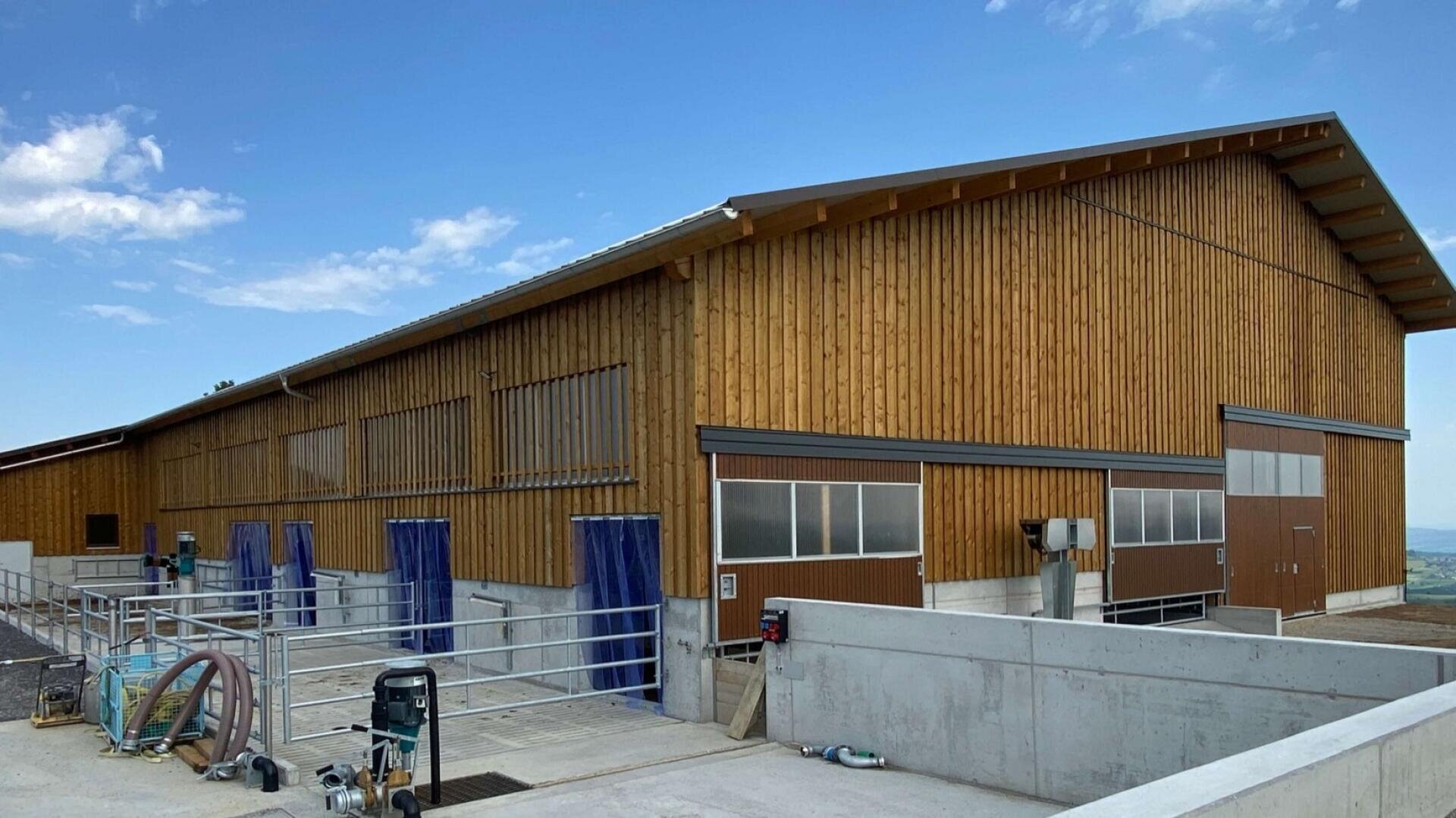 Eine große hölzerne Scheune mit schrägem Dach, braunen Türen und blauen Vorhängen an den Eingängen, umgeben von Betonbarrieren und verschiedenen Geräten unter einem klaren blauen Himmel.