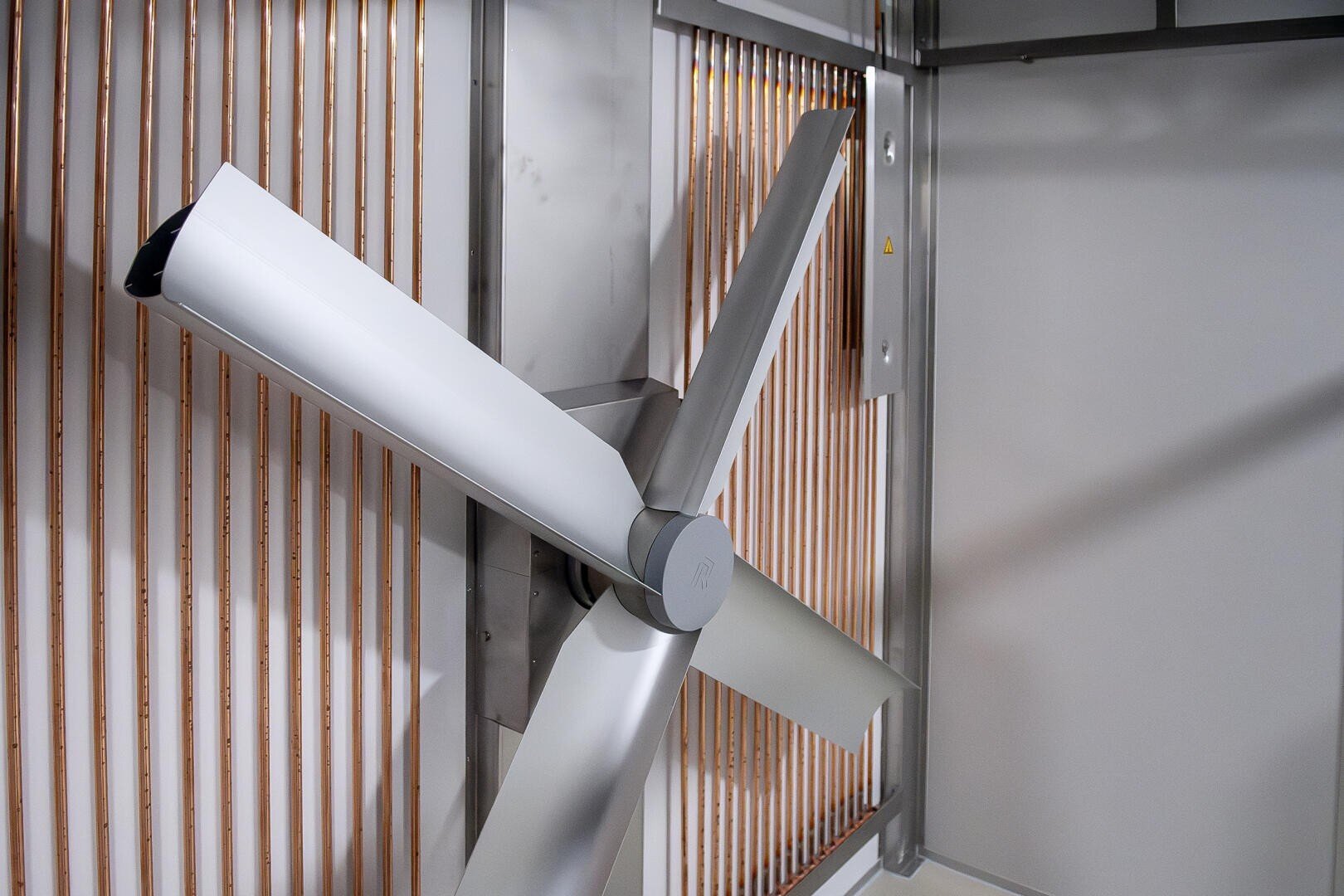 Ein großer, moderner industrieller Deckenventilator mit drei silbernen Flügeln auf einem Metallrahmen vor einem Hintergrund aus vertikalen Kupferrohren und einer weißen Wand.