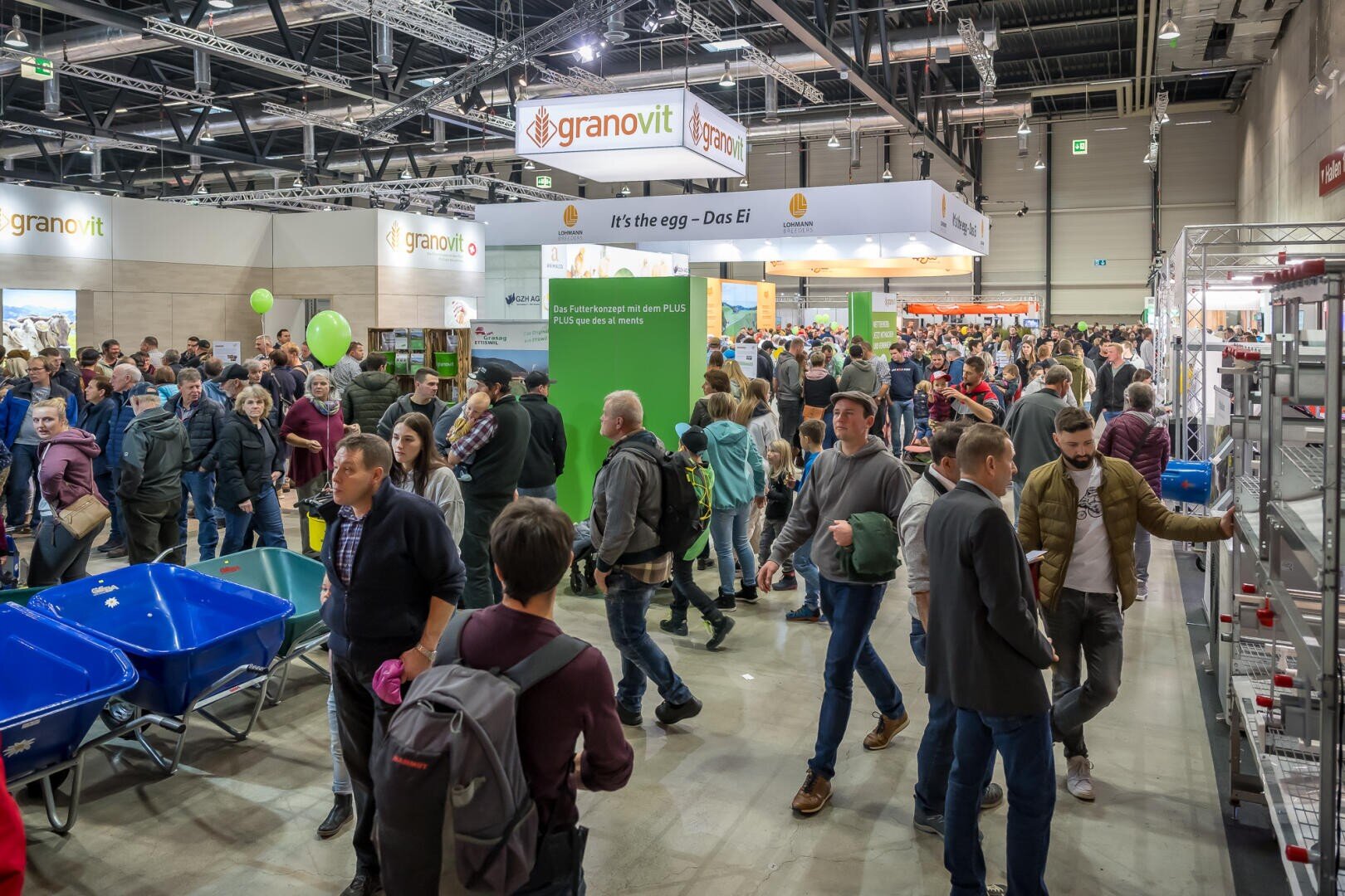 Eine belebte Messe in einer Halle mit vielen Menschen, die um die verschiedenen Stände und Auslagen herumgehen und interagieren, darunter blaue Schubkarren und grüne Schilder unter hellen Oberlichtern. Das granovit-Logo ist über einigen Ständen zu sehen.