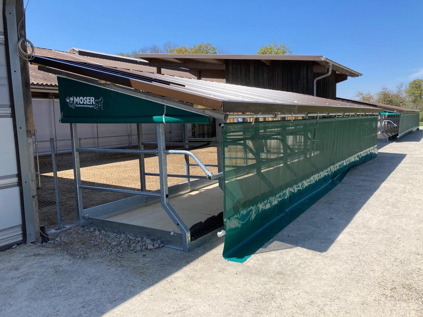 Ein Viehstall aus Metall und grünem Stoff mit dem Moser-Branding steht neben einer Scheune unter einem klaren blauen Himmel. Der Unterstand hat ein schräges Metalldach und ist auf der dem Gehweg zugewandten Seite offen.