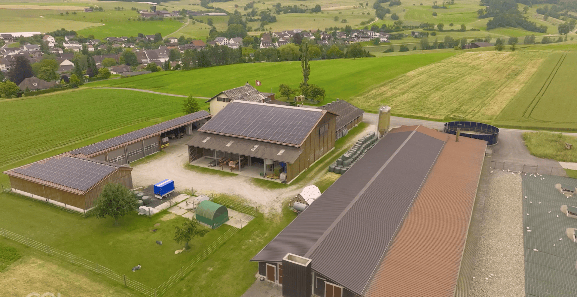 Luftaufnahme eines Bauernhofs mit mehreren Gebäuden, viele davon mit Sonnenkollektoren auf den Dächern, umgeben von grünen Feldern und einem entfernten Dorf unter einem bewölkten Himmel.