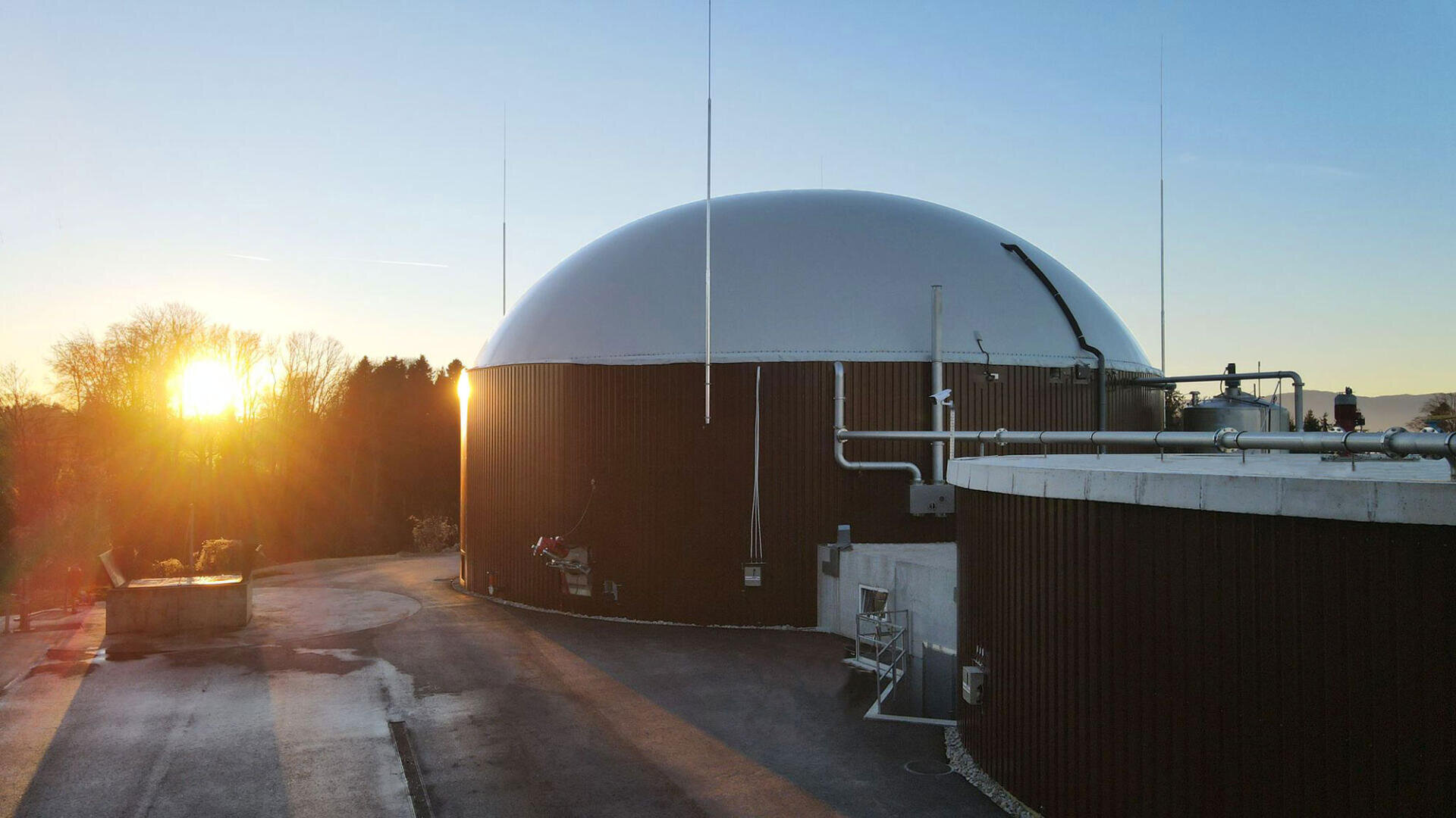 Eine große, kuppelförmige Biogasanlage mit Rohren und Tanks steht bei Sonnenaufgang unter einem klaren Himmel, umgeben von Bäumen und einer leeren, gepflasterten Fläche.