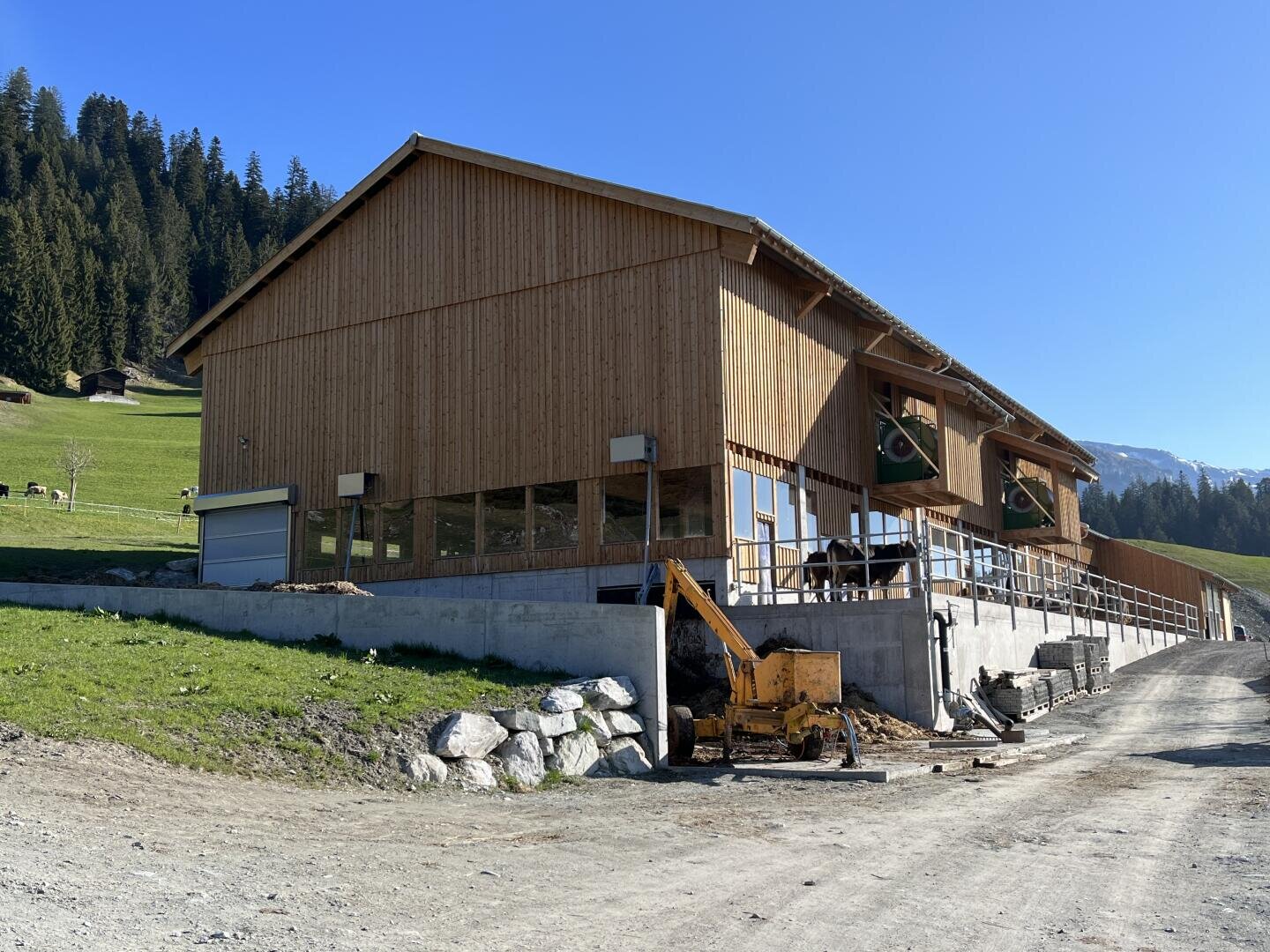 Eine große Holzscheune mit breiten Fenstern steht auf einem Betonfundament neben einer Schotterstraße, mit grünen Hügeln und Bäumen im Hintergrund unter einem klaren blauen Himmel. Davor ist ein gelber Bagger geparkt.