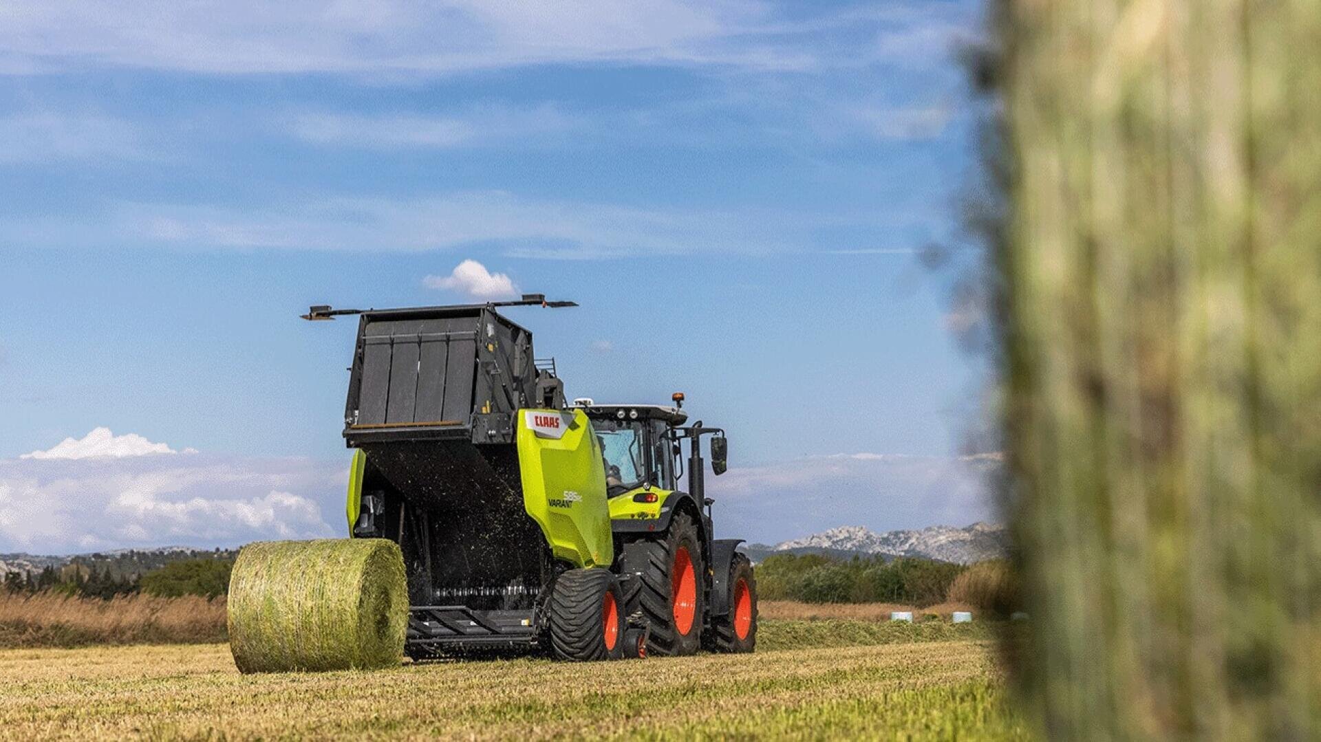 Ein grüner Traktor mit roten Rädern presst und entlässt einen großen Heuballen auf ein grasbewachsenes Feld unter blauem Himmel; ein weiterer Heuballen ist im Vordergrund zu sehen.