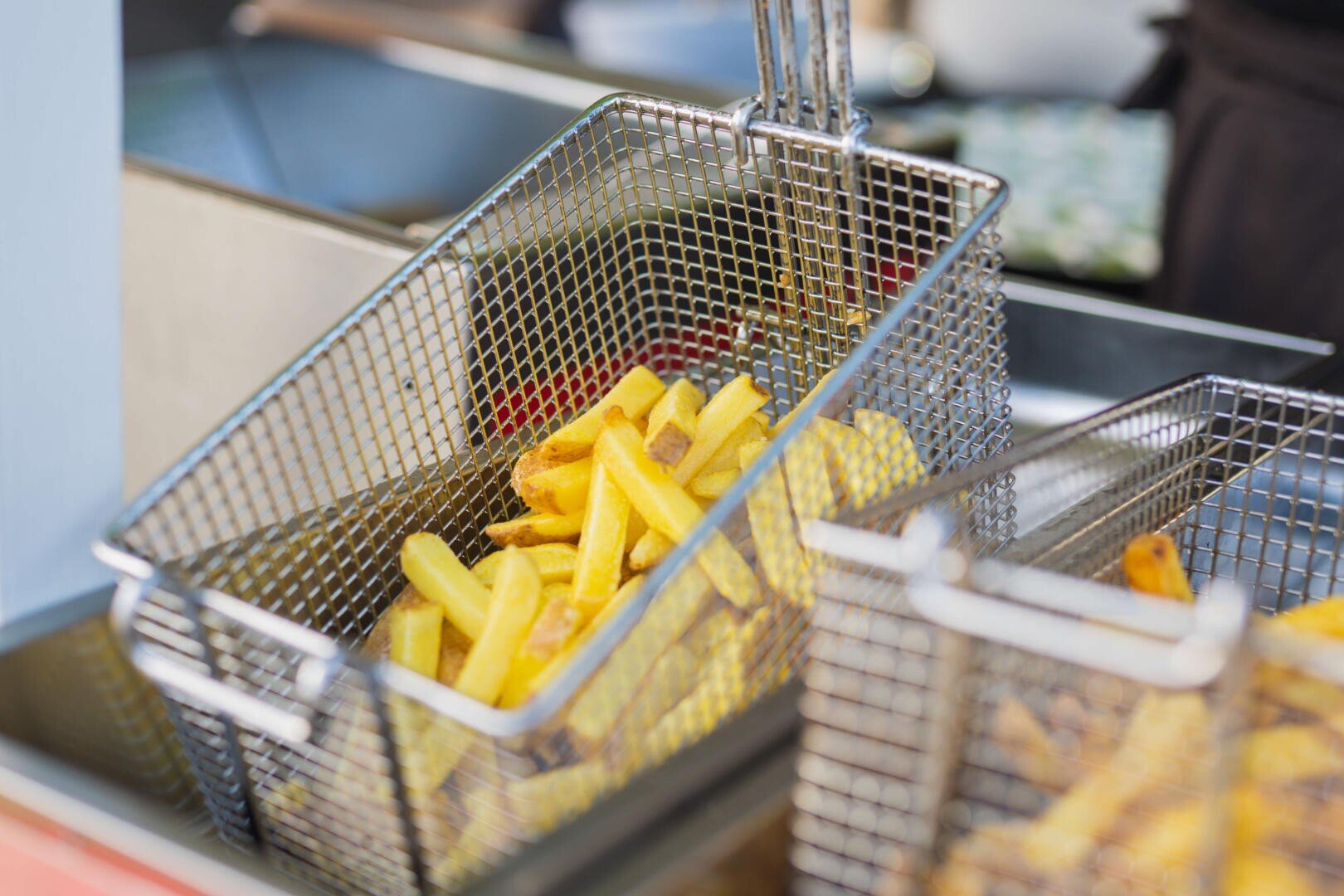 Ein mit frisch zubereiteten Pommes frites gefüllter Metallkorb wird aus einer Fritteuse gehoben, ein weiterer Korb ist im Vordergrund zu sehen.