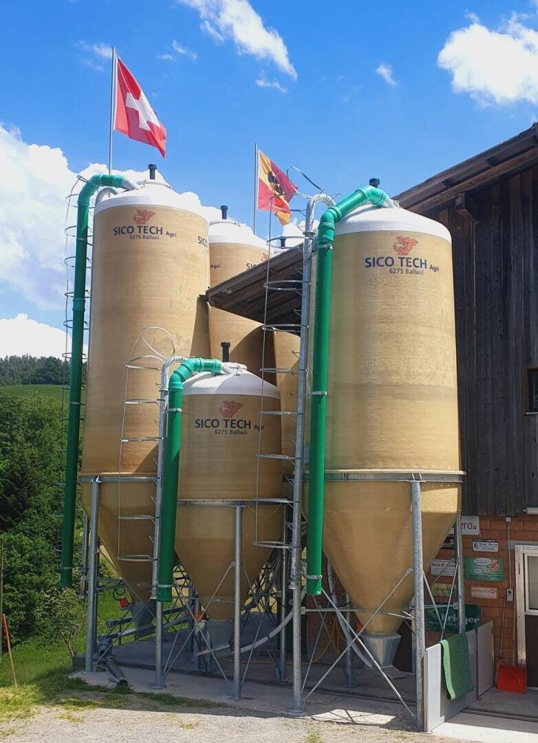 Vier große hellbraune Sico-Tech-Silos mit grünen Rohren stehen neben einem Holzgebäude. Schweizer und regionale Flaggen sind auf dem Dach. Der Himmel ist blau mit Wolken, und im Hintergrund ist Grün zu sehen.
