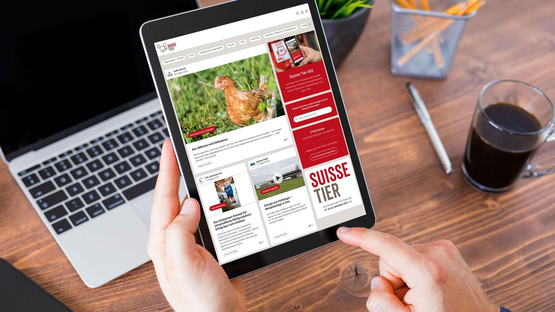 Eine Person hält und benutzt ein Tablet, auf dem eine Website mit Artikeln angezeigt wird, darunter ein Bild eines Huhns. Das Tablet steht auf einem Holztisch neben einem Laptop, einem Glas Kaffee und einem Stifthalter.