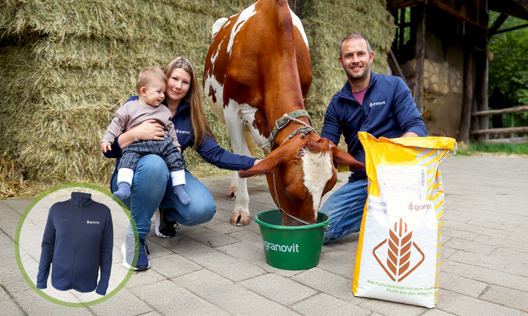 Ein lächelnder Mann und eine Frau mit einem Kind posieren neben einer braun-weißen Kuh, die aus einem grünen Eimer frisst. Ein großer Sack mit Tierfutter und ein Bild einer blauen Jacke sind ebenfalls zu sehen. Sie stehen draußen in der Nähe von Heuballen.