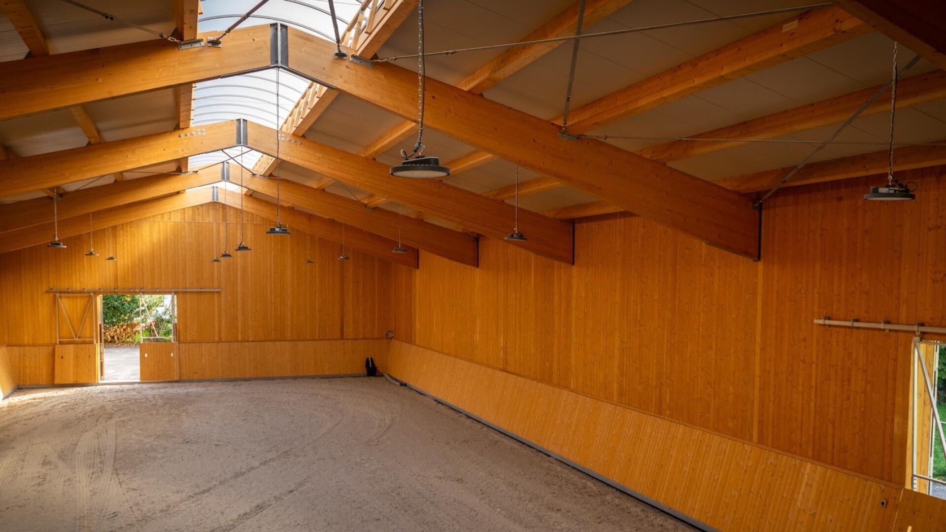 Große Reithalle mit Holzwänden, Sichtbalken, Oberlichtern und Hängelampen. Der mit Sand bedeckte Boden ist nach links gewölbt, und das Sonnenlicht fällt durch eine offene Tür und ein Fenster auf der anderen Seite.