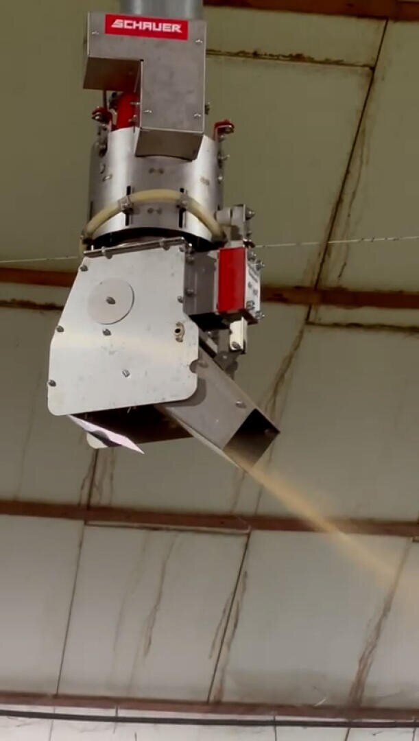 Ein automatischer Futterautomat aus Metall mit der Aufschrift Schauer hängt von der Decke und gibt Futter in einem kontrollierten Strom in einem Stall oder einer landwirtschaftlichen Einrichtung aus.