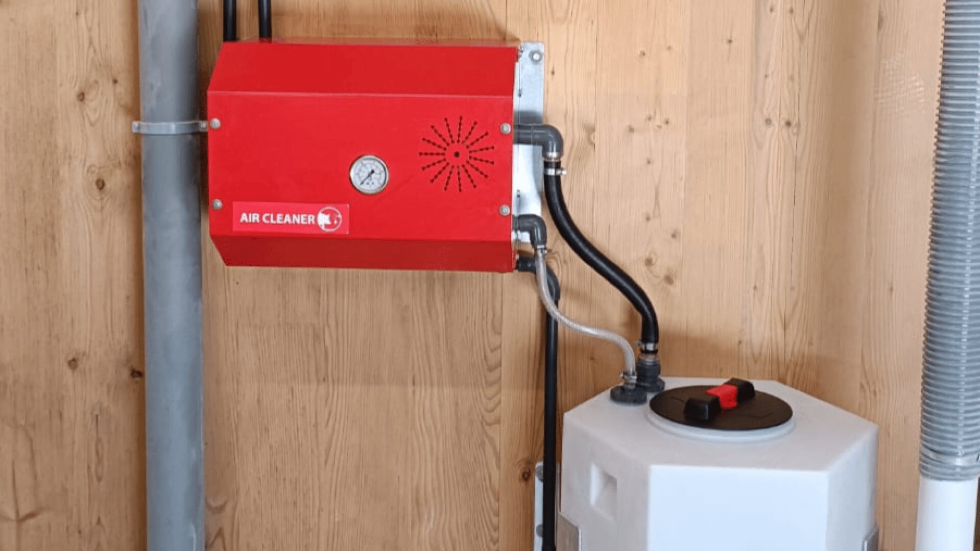 Ein roter Luftreiniger ist an einer Holzwand montiert und durch schwarze Schläuche mit einem weißen Behälter mit schwarzem und rotem Deckel neben einem grauen Rohr verbunden.