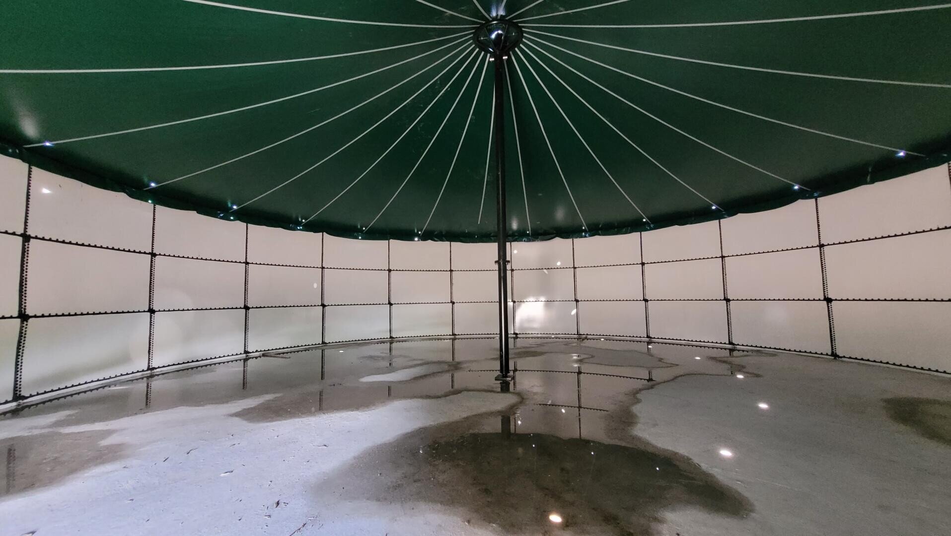 Das Innere eines großen, runden Wassertanks mit einem grünen Kuppeldach, das von einem zentralen Pfosten und radialen Balken getragen wird. Auf dem Betonboden sind Spiegelungen und Wasserflecken zu sehen. Die Wände des Tanks sind hell gefärbt.