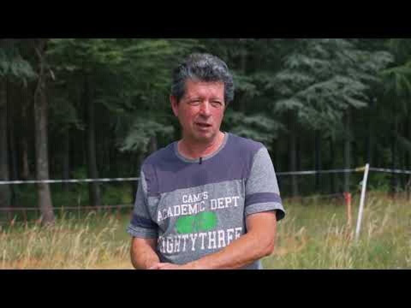 Ein Mann mittleren Alters mit ergrauendem Haar steht im Freien vor einem Waldgebiet und trägt ein lässiges graues T-Shirt mit aufgedrucktem Text. Der Hintergrund ist grasbewachsen mit Bäumen und einigen Zäunen.