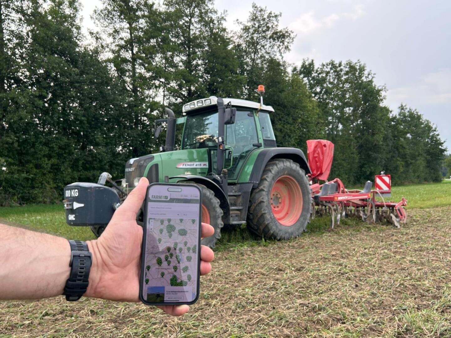 Eine Person hält ein Smartphone, auf dem eine Landwirtschafts-App mit einer Karte angezeigt wird, vor einem grünen Traktor und roten landwirtschaftlichen Geräten auf einem Feld, mit Bäumen und bewölktem Himmel im Hintergrund.