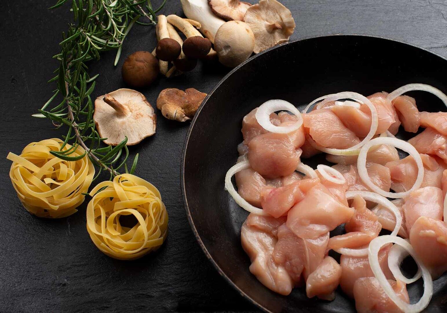 Rohes gewürfeltes Hühnerfleisch und in Scheiben geschnittene Zwiebeln in einer Pfanne, daneben auf einer dunklen Fläche ungekochte Tagliatelle-Nester, frischer Rosmarin und verschiedene Pilze.