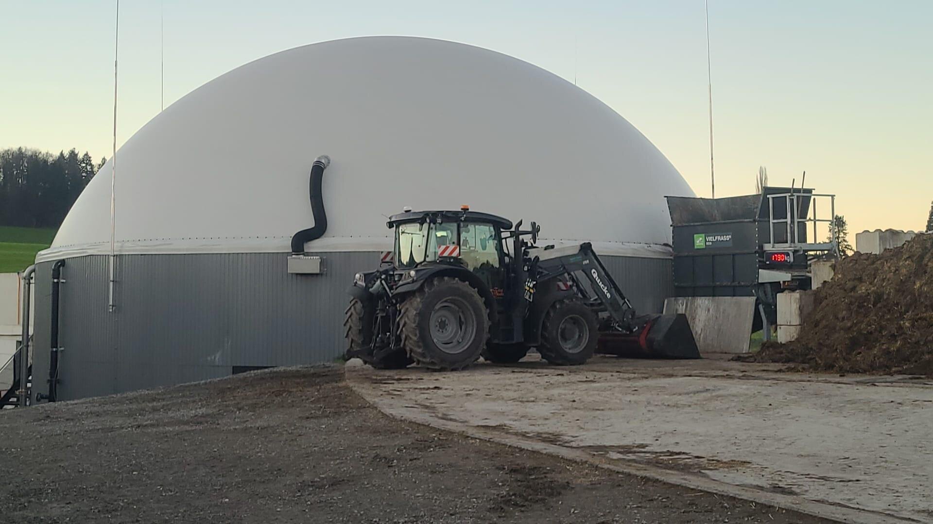 Ein schwarzer Traktor parkt in der Nähe einer großen, kuppelförmigen Biogasanlage mit grauem Sockel und weißem Dach, umgeben von Ackerland und Erdhaufen auf einem Bauernhof.