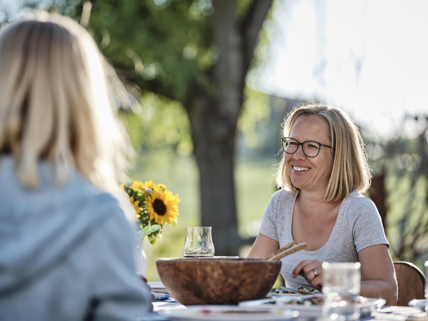 Zwei Frauen sitzen an einem Tisch im Freien, unterhalten sich und lächeln. Die eine Frau trägt eine Brille und ein graues Hemd. Auf dem Tisch stehen eine hölzerne Salatschüssel, ein Glas und eine Vase mit Sonnenblumen, und das Sonnenlicht fällt durch die Bäume.