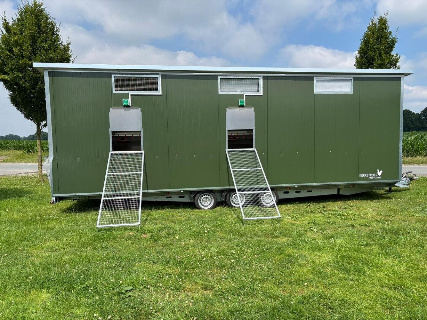 Ein grüner mobiler Hühnerstall mit drei Metallgitterrampen, die zu kleinen Türen führen, steht auf einem grasbewachsenen Feld mit Bäumen und einem teilweise bewölkten Himmel im Hintergrund.